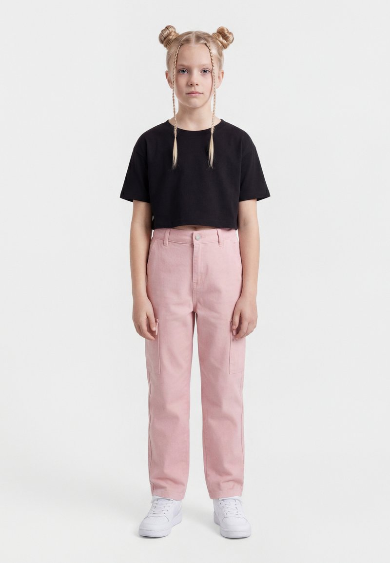 Fiatal lány szőke hajjal, két kontyban és fonatokkal, fekete, rövid felsőt, rózsaszín nadrágot és fehér tornacipőt visel, előre nézve áll.