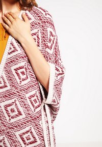 Strikket cardigan med et vinrødt og hvidt geometrisk mønster. Åben front, brede ærmer og en tekstureret finish. Varm, afslappet stil.