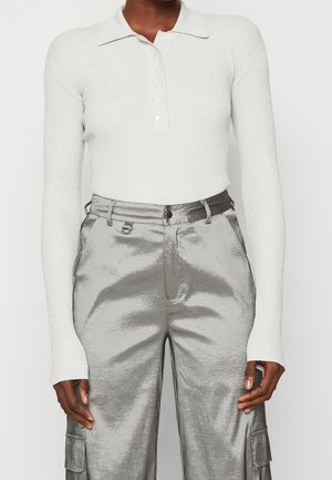 Personne portant un polo blanc côtelé à manches longues, rentré dans un pantalon cargo en satin argenté taille haute avec poches latérales.