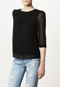 Svart genomskinlig blus med trekvartsärmar, plisserad frontdetalj och lagerdesign; kombinerad med slitna ljusblå jeans.