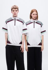 Λευκό πουκάμισο με φερμουάρ μέχρι τη μέση, με μοτίβο στους ώμους σε μέντα και μπορντό. Διαθέτει χαλαρή εφαρμογή, ελαστικό κάτω μέρος και πλαϊνές τσέπες. Μαύρο παντελόνι.