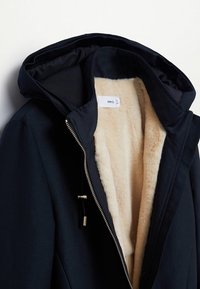 Schwarze Außenjacke mit einem plüschigen beigen Innenfutter, ausgestattet mit Kapuze, sichtbarem Reißverschluss und Kordelzugdetails. Weicher Stoff und modernes Design.