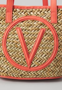Πλεκτή τσάντα από άχυρο με κοραλλί δερμάτινες λεπτομέρειες, με κυκλικό λογότυπο που έχει ένα 'V' στο κέντρο και επίπερους χειροκροτητές.