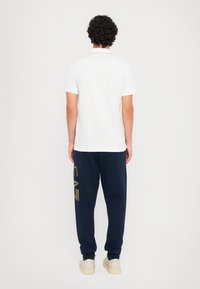 Balti trumpos rankovės marškinėliai ir tamsiai mėlyni treningai su auksinėmis raidėmis, derinami su kreminiais sportbačiais. Paprastas, kasdieniškas aprangos komplektas be matomų raštų.