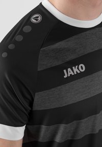 Maglietta atletica nera con motivo a righe grigie, dotata di un colletto bianco e dettagli sulle spalle. Include un logo "JAKO" in un colore a contrasto.