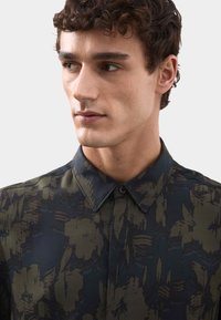 Kurzarmhemd, dunkelgrün mit floralem Muster, gestreifter Kragen, Knopfverschluss, aus strukturiertem Stoff. Detailreiche florale Akzente sichtbar.