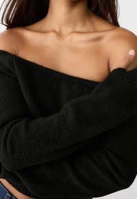 Pull noir épaules dénudées en tissu doux et texturé avec manches longues, mettant en valeur une coupe décontractée et des poignets côtelés.