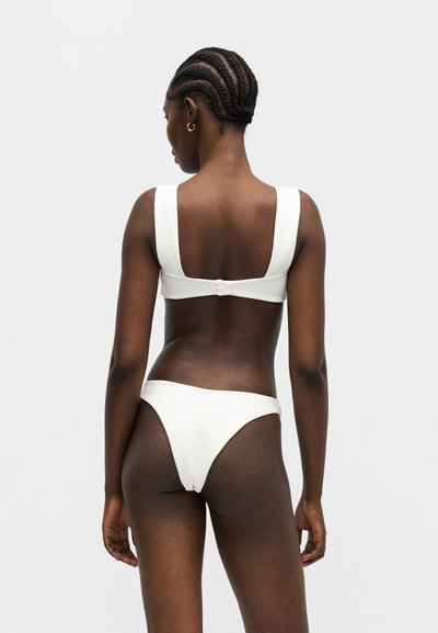 Γυναίκα με κοτσιδάκια, που φοράει λευκό μπικίνι, φωτογραφημένη από πίσω σε μονόχρωμο γκρι φόντο.