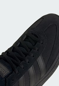 Czarny but sportowy z cholewką z siateczki i zamszu, okrągłymi sznurowadłami oraz trzema poziomymi paskami po boku. Podeszwa z gumy z teksturowanym bieżnikiem.