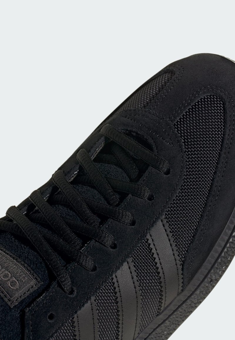 Zapatilla deportiva negra con parte superior de malla y gamuza, cordones redondos y tres franjas horizontales en el lateral. Suela de goma con agarre texturizado.