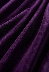 Tejido de terciopelo en un tono púrpura profundo, mostrando una textura suave y un caída fluida con un brillo sutil. La superficie presenta ligeras variaciones que añaden profundidad.