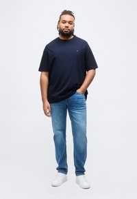 Άνδρας με πλεξούδες φοράει μπλε σκούρο t-shirt, ανοιχτόχρωμο τζιν και λευκά αθλητικά παπούτσια, στέκεται με το ένα χέρι στην τσέπη σε λευκό φόντο.