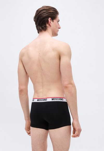 Άνδρας με γυμνή πλάτη, φοράει μαύρα μπόξερ Moschino με λευκή ζώνη μέσης, στέκεται μπροστά σε απλό ανοιχτό γκρι φόντο.