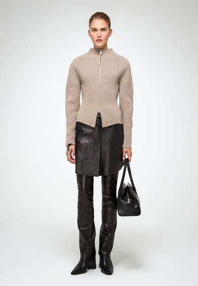 Beige bordázott pulóver magas gallérral és ezüst cipzárral, fekete bőrszoknyával és testhezálló fekete nadrággal párosítva. Fekete kézitáska is tartozik hozzá.
