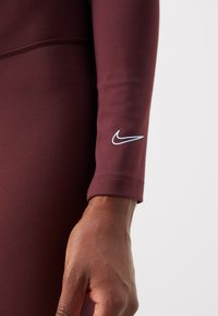 Burgundin värinen venytettävä urheilupaita pitkähihaisella mallilla, istuva muotoilu, litteät saumat ja valkoinen Nike-logo hiassa. Silkkinen pinta.