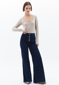 Béžový dlouhý rukáv top s čtvercovým výstřihem spárovaný s tmavě modrými širokými kalhotami s vysokým pasem a zapínáním na pět knoflíků.