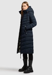 Jūros mėlynumo pūkų striukė su ilgu, tiesiu siluetu, horizontaliai siuvinėtu raštu, reguliuojama kapuce ir priekiniu užtrauktuku, derinama su juodais batų raišteliais.