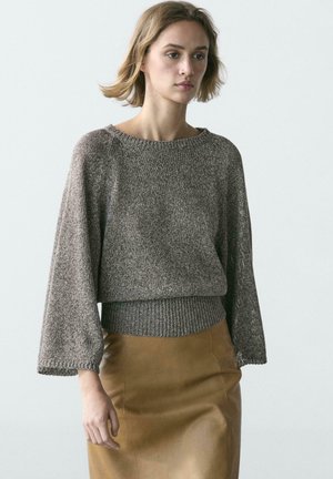 Femme portant un pull en maille gris ample à larges manches et une jupe marron ajustée, debout devant un fond clair uni.