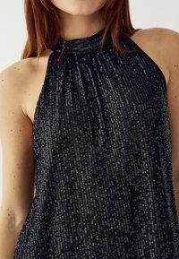 Čierny halter top vyrobený z flitrovanej tkaniny, s textúrovaným povrchom a voľným strihom, odrážajúci svetlo v rôznych vzoroch.