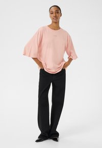 Heled roosa oversized t-särk lühikeste, laiade varrukatega ning diskreetse logo detailiga, kombineeritud kõrge vöökohaga mustade laia säärega teksade ja mustade terava nina kingadega.