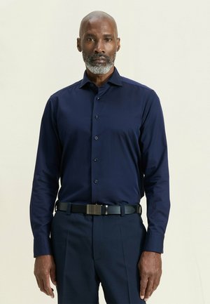 Man met grijze baard die een donkerblauw overhemd met knopen en bijpassende donkerblauwe broek met zwarte riem draagt, staand tegen een lichte achtergrond.