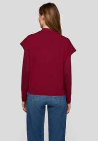 Bordeauxroter Pullover mit kurzen, strukturierten Ärmeln, hohem Kragen und kurzem Rückensaum. Kombiniert mit blauen, locker sitzenden Jeans.