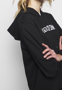 Černá mikina s klokaní kapsou, kapucí na šňůrku a vytištěným bílým logem přes hrudník. Měkká bavlněná tkanina s žebrovanými manžetami.