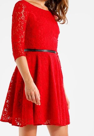 Femme portant une robe en dentelle rouge longue jusqu'aux genoux, manches trois-quarts et ceinture noire cloutée à la taille.