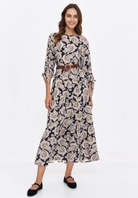 Woman smiling, wearing a long black dress with beige paisley print, brown belt, and black flat shoes, standing against a white background.