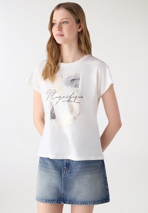 Giovane donna che indossa una maglietta bianca con disegno di piuma nera e dorata e testo "Magnifique", abbinata a una gonna di jeans blu, in piedi con le mani dietro la schiena.