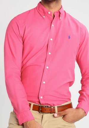 Pink button-down cotton shirt with long sleeves, white buttons, and a blue logo on the chest; worn with a brown belt and beige pants.