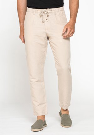 Uomo che indossa pantaloni beige con coulisse e scarpe slip-on verde oliva, in piedi davanti a uno sfondo bianco.