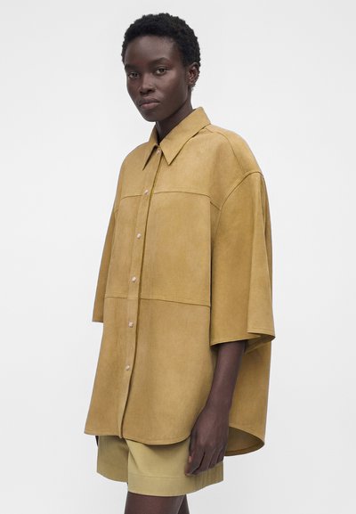 Γυναίκα φοράει υπερμεγέθη μπεζ πουκάμισο από σουέντ με κουμπιά και μανίκια μέχρι τον αγκώνα, καθώς και ταιριαστά σορτς, σε απλό λευκό φόντο.