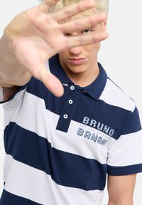 Ung man med blont hår som bär en marinblå och vitrandig polo tröja och räcker upp handen mot kameran med ett allvarligt uttryck.