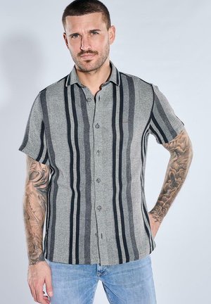 Mann mit kurzen Haaren und Tätowierungen an beiden Armen trägt ein graues Hemd mit schwarzen vertikalen Streifen und hellblaue Jeans, steht vor einem einfarbigen Hintergrund.