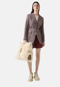 Blazer z vzorované tkaniny s páskem v pase, nošený přes krémový top a vínové šortky. Drží huňatou krémovou tašku a má na sobě béžové vysoké boty.