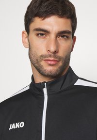 Schwarze Sportjacke mit weißen Akzenten, hohem Kragen, durchgehendem Reißverschluss und "JAKO"-Logo auf der linken Brust. Glatte Textur, sportliches Design.