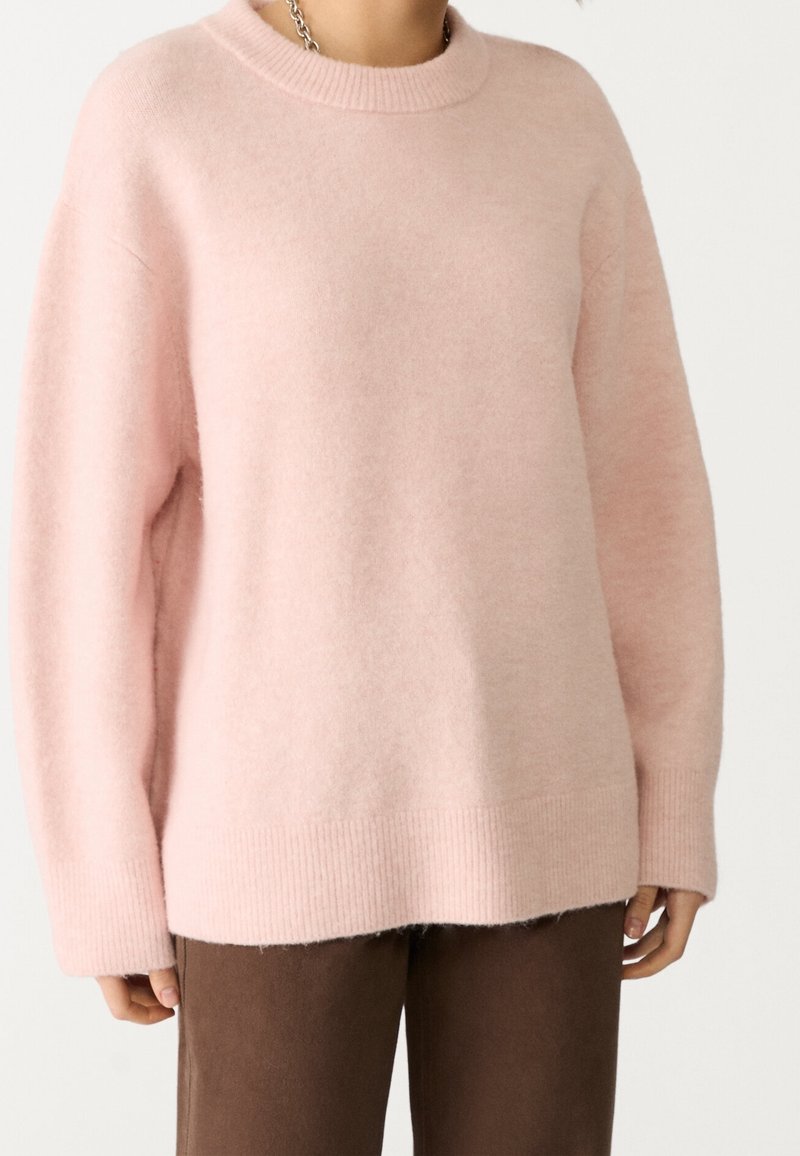 Pull en maille rose clair avec un col rond, un ourlet côtelé et une coupe ample ; porté avec un pantalon marron. Texture douce, manches longues.