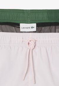 Costumi da bagno Lacoste con un corpo rosa chiaro, cintura verde e cordoncino bianco. Presentano una fodera in rete con l'etichetta del logo Lacoste visibile.