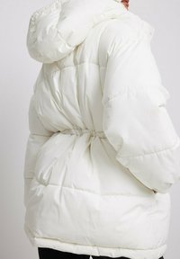 Chaqueta acolchada blanca con capucha, que presenta una cintura ajustada, diseño acolchado y textura suave. Ofrece un aspecto voluminoso y cálido.