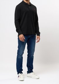 Polo shirt nero a maniche lunghe con colletto e logo rosso, abbinato a jeans blu e sneakers bianche con accenti a righe.