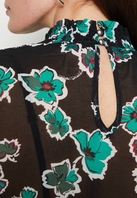 Schwarze transparente Bluse mit buntem Blumenmuster in Blaugrün und Weiß, gerafftem Ausschnitt, geknöpftem Schlüsselloch-Detail im Rücken mit zwei dekorativen Knöpfen.