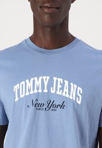 Gaiši zils kokvilnas t-krekls ar baltu tekstu "TOMMY JEANS" un melnu "New York since 1985", apaļa kakla dizains, īsas piedurknes.