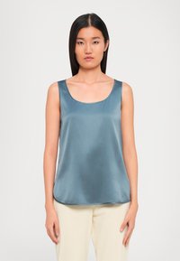 Woman wearing sleeveless shiny steel blue top and cream pants, standing against a plain white background, facing forward.