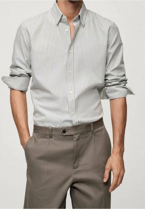 Man draagt een lichtgrijs-wit gestreept overhemd met verticale strepen, mouwen opgerold, en grijze nette broek, handen in en bij de zakken.