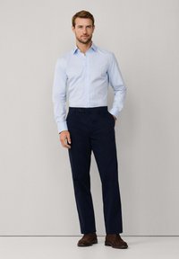 Man staat met een lichtblauw geblokt overhemd, marineblauwe broek en bruine schoenen tegen een effen lichtgrijze achtergrond.