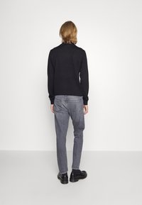 Person med axellångt hår iförd en svart långärmad tröja, grå jeans och svarta skor, stående vänd mot en vit vägg.