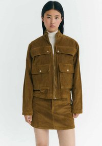 Brown corduroy jacket with zip closure, two front pockets, and matching skirt. Features vertical ribbing and a structured design.