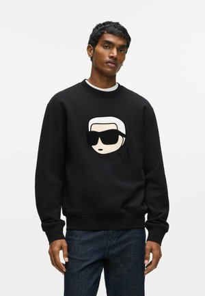 Jeune homme aux cheveux courts et noirs portant un sweat-shirt noir avec un visage de dessin animé portant des lunettes de soleil et un jean bleu sur un fond uni.