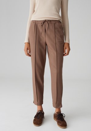 Pantaloni marroni su misura con vita con coulisse, pieghe laterali e gambe affusolate, abbinati a sneaker marroni scuri. Tessuto dalla texture liscia.